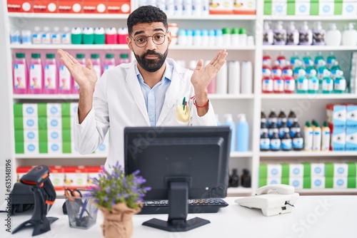 Hispanic man with beard working at pharmacy drugstore clueless and confused expression with arms and hands raised. doubt concept.