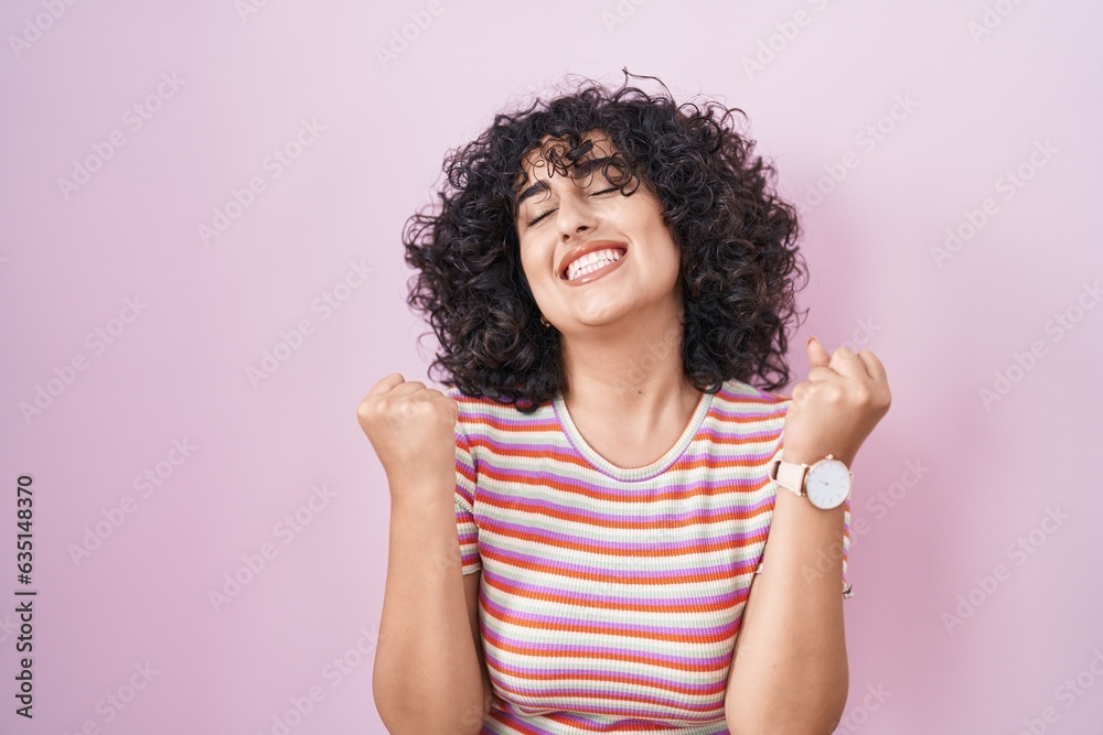 Young middle east woman standing over pink background celebrating surprised and amazed for success with arms raised and eyes closed. winner concept. Young middle east woman standing over pink background celebrating surprised and amazed for success with arms raised and eyes closed. winner concept.