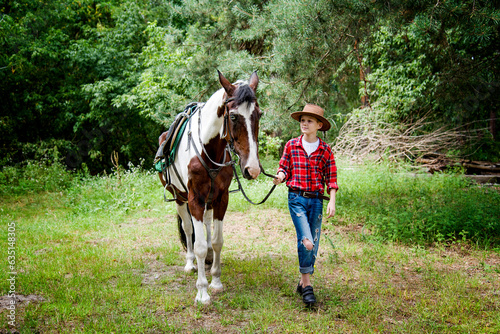 little handsome blonde smiling boy in red checkered shirt and cowboy brown hat walking and hugging horse in green forest on sunny day 