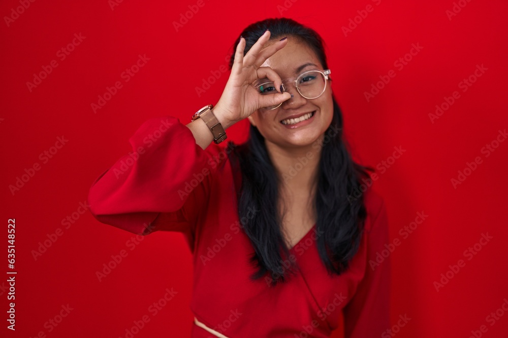 Asian young woman standing over red background doing ok gesture with hand smiling, eye looking through fingers with happy face. Asian young woman standing over red background doing ok gesture with hand smiling, eye looking through fingers with happy face.