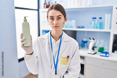 Young caucasian woman working at scientist laboratory holding body lotion thinking attitude and sober expression looking self confident