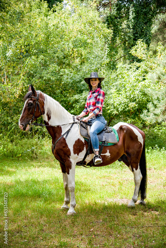 beautiful smiling brunette cowboy woman in hat and red checkered shirt riding horse in green forest  © Tetatet