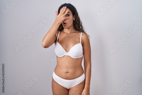 Young hispanic woman wearing white lingerie peeking in shock covering face and eyes with hand, looking through fingers with embarrassed expression. © Krakenimages.com