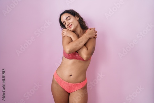 Canvas-taulu Young hispanic woman wearing lingerie over pink background hugging oneself happy and positive, smiling confident