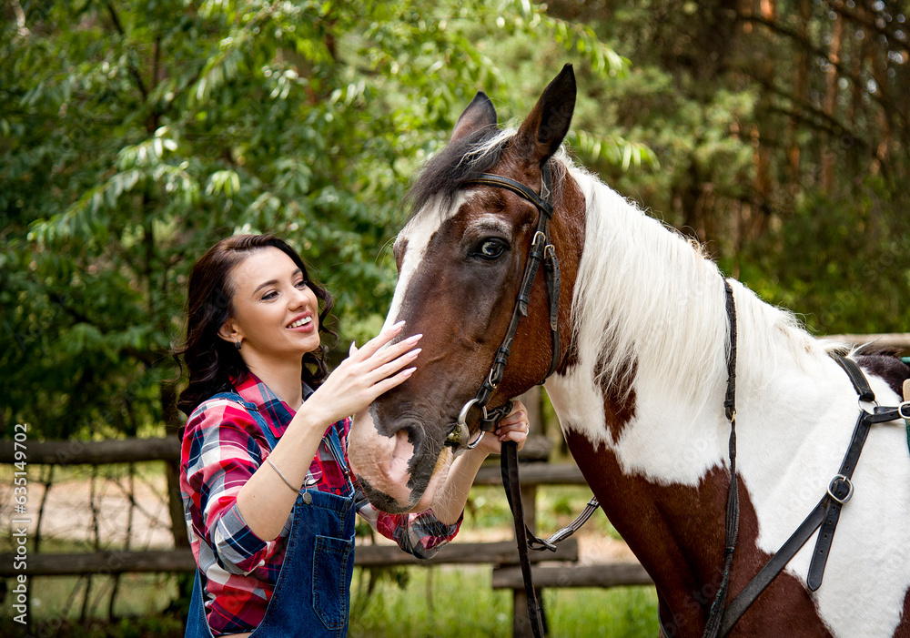 Obraz premium portrait of beautiful smiling brunette cowboy woman in red checkered shirt walking and hugging with horse in green forest on sunny day
