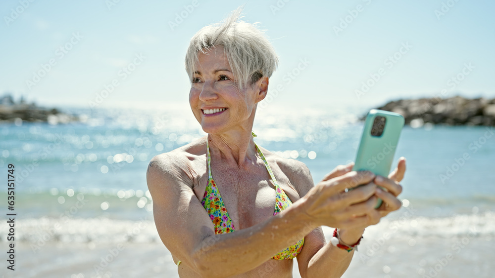 Middle age blonde woman tourist wearing bikini using smartphone at the beach