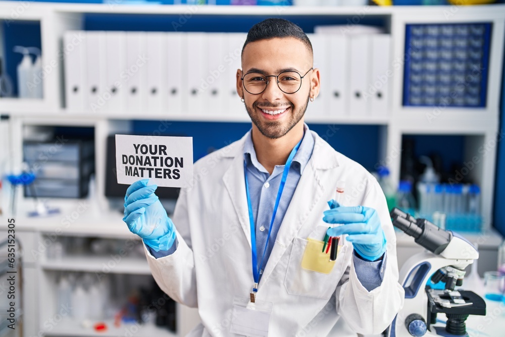 Young hispanic man working at scientist laboratory holding your donation matters holding blood sample smiling with a happy and cool smile on face. showing teeth. Young hispanic man working at scientist laboratory holding your donation matters holding blood sample smiling with a happy and cool smile on face. showing teeth.