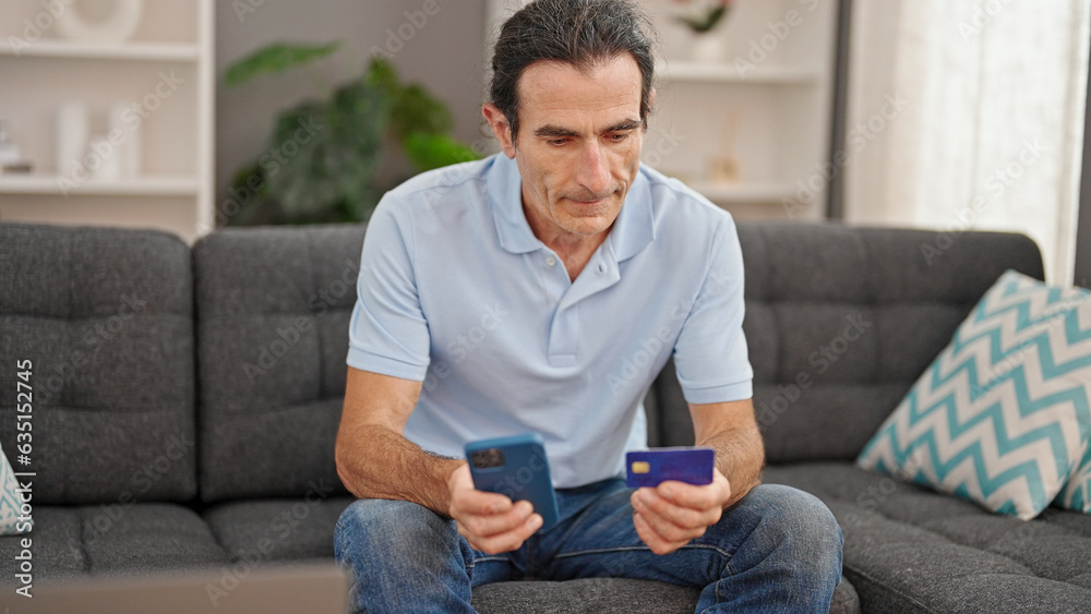 Middle age man shopping with smartphone and credit card sitting on sofa at home