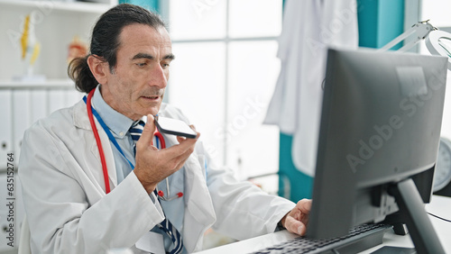 Middle age man doctor using computer sending voice message by smartphone at the clinic