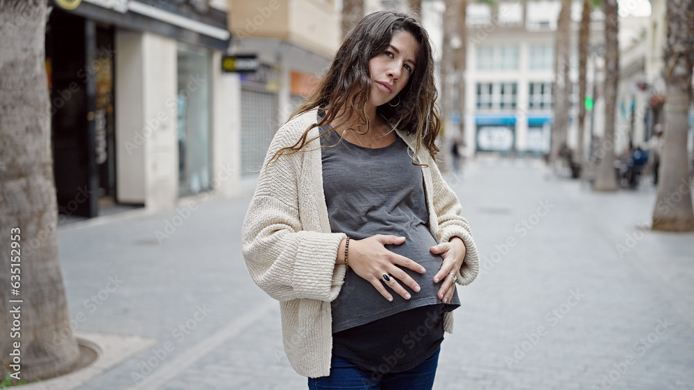 Fototapeta premium Young pregnant woman standing with serious expression touching belly at street