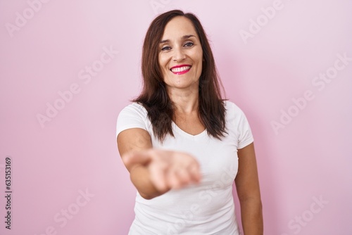 Middle age brunette woman standing over pink background smiling cheerful offering palm hand giving assistance and acceptance.