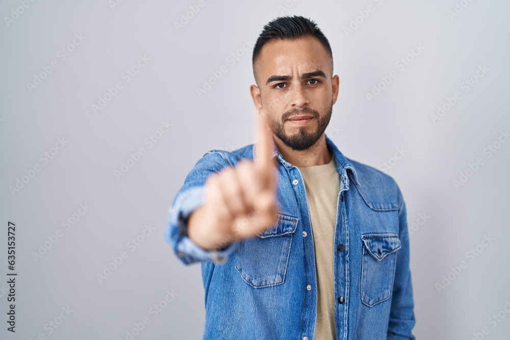 Young hispanic man standing over isolated background pointing with finger up and angry expression, showing no gesture Young hispanic man standing over isolated background pointing with finger up and angry expression, showing no gesture