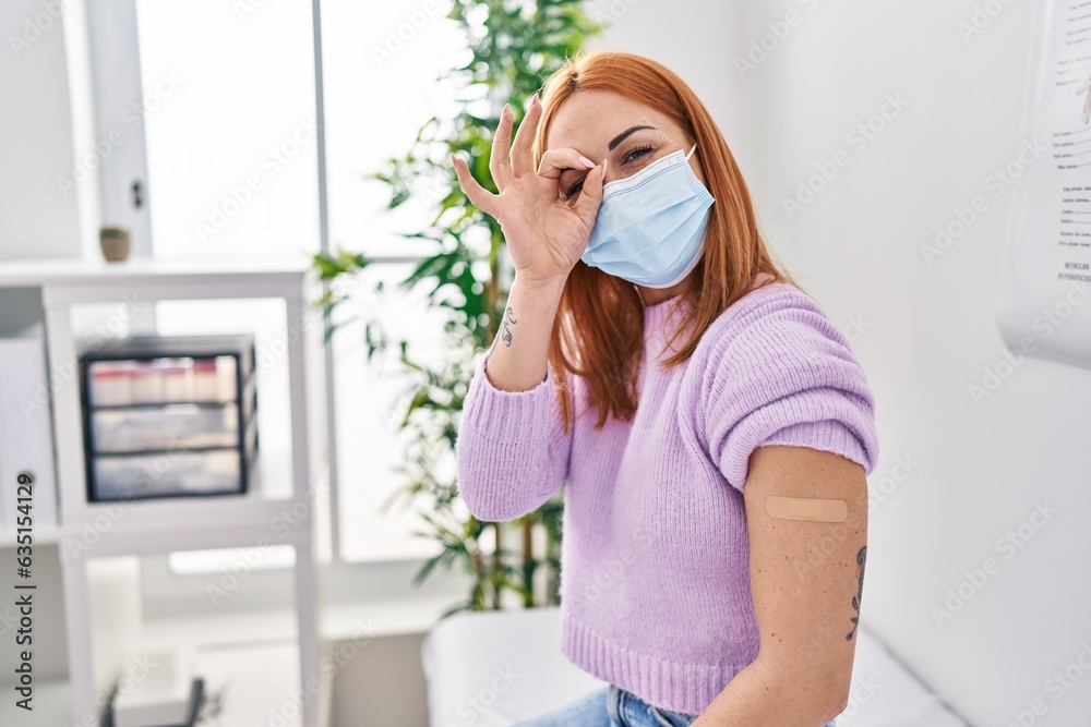 Young woman getting vaccine showing arm with band aid smiling happy doing ok sign with hand on eye looking through fingers Young woman getting vaccine showing arm with band aid smiling happy doing ok sign with hand on eye looking through fingers