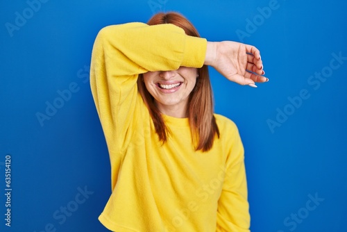 Young woman standing over blue background covering eyes with arm smiling cheerful and funny. blind concept. © Krakenimages.com