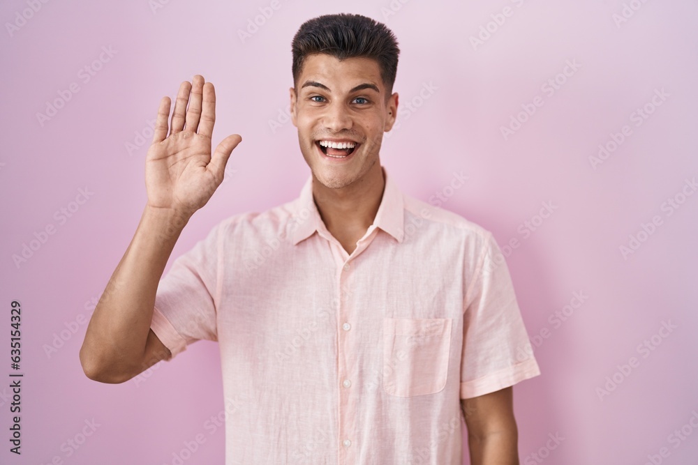 Young hispanic man standing over pink background waiving saying hello happy and smiling, friendly welcome gesture Young hispanic man standing over pink background waiving saying hello happy and smiling, friendly welcome gesture