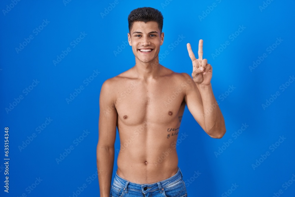 Young hispanic man standing shirtless over blue background showing and pointing up with fingers number two while smiling confident and happy.
