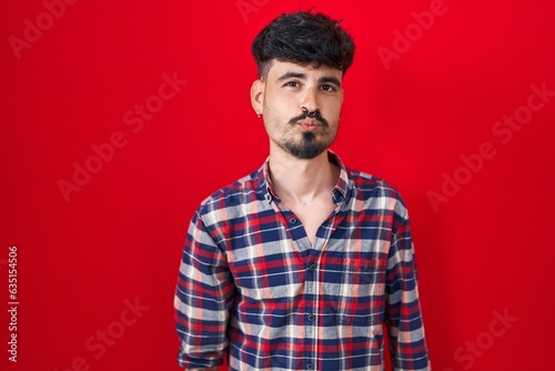 Young hispanic man with beard standing over red background looking at the camera blowing a kiss on air being lovely and sexy. love expression.