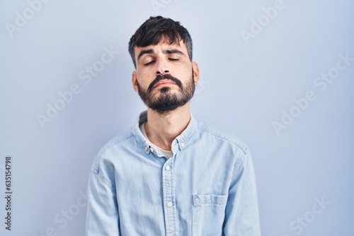 Young hispanic man with beard standing over blue background looking sleepy and tired, exhausted for fatigue and hangover, lazy eyes in the morning.