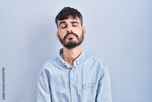 Young hispanic man with beard standing over blue background looking sleepy and tired, exhausted for fatigue and hangover, lazy eyes in the morning. © Krakenimages.com