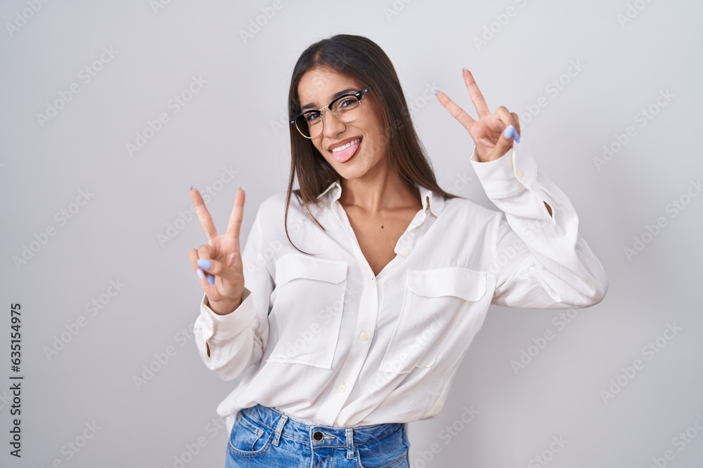 Young brunette woman wearing glasses smiling with tongue out showing fingers of both hands doing victory sign. number two. Young brunette woman wearing glasses smiling with tongue out showing fingers of both hands doing victory sign. number two.