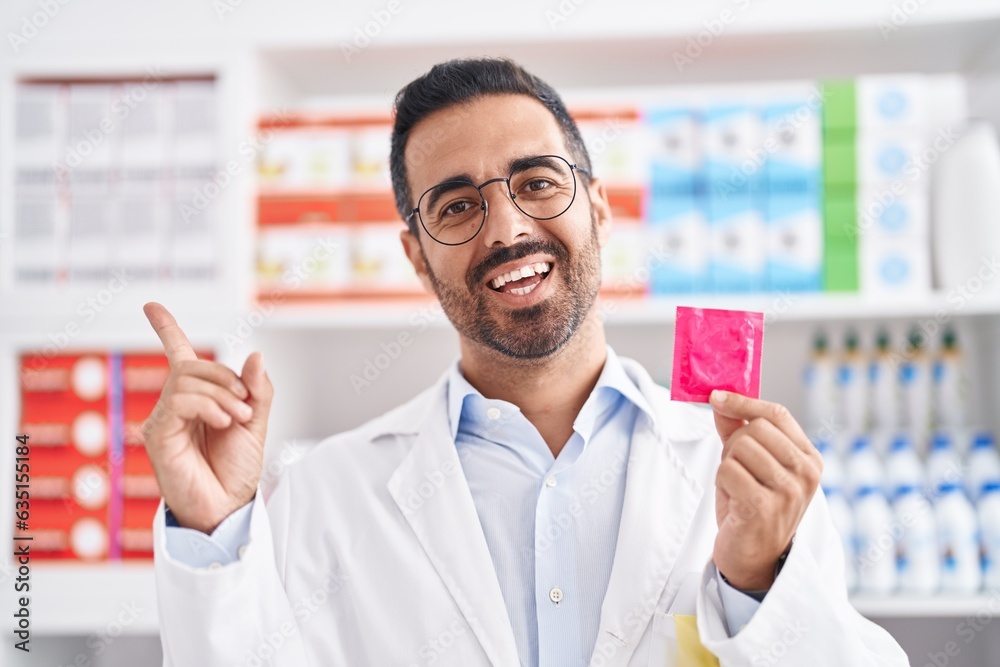 Hispanic man with beard working at pharmacy drugstore holding condom smiling happy pointing with hand and finger to the side Hispanic man with beard working at pharmacy drugstore holding condom smiling happy pointing with hand and finger to the side