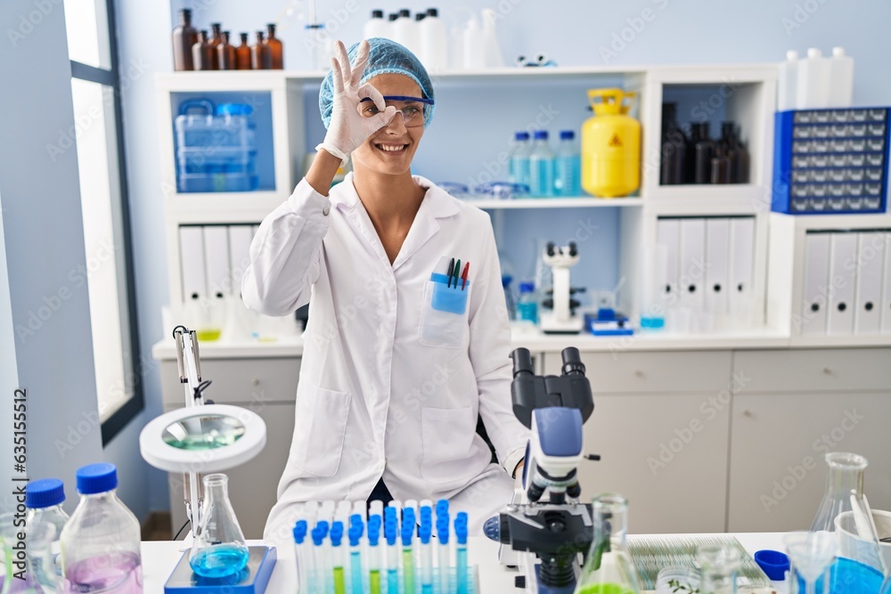 Brunette woman working at scientist laboratory doing ok gesture with hand smiling, eye looking through fingers with happy face.