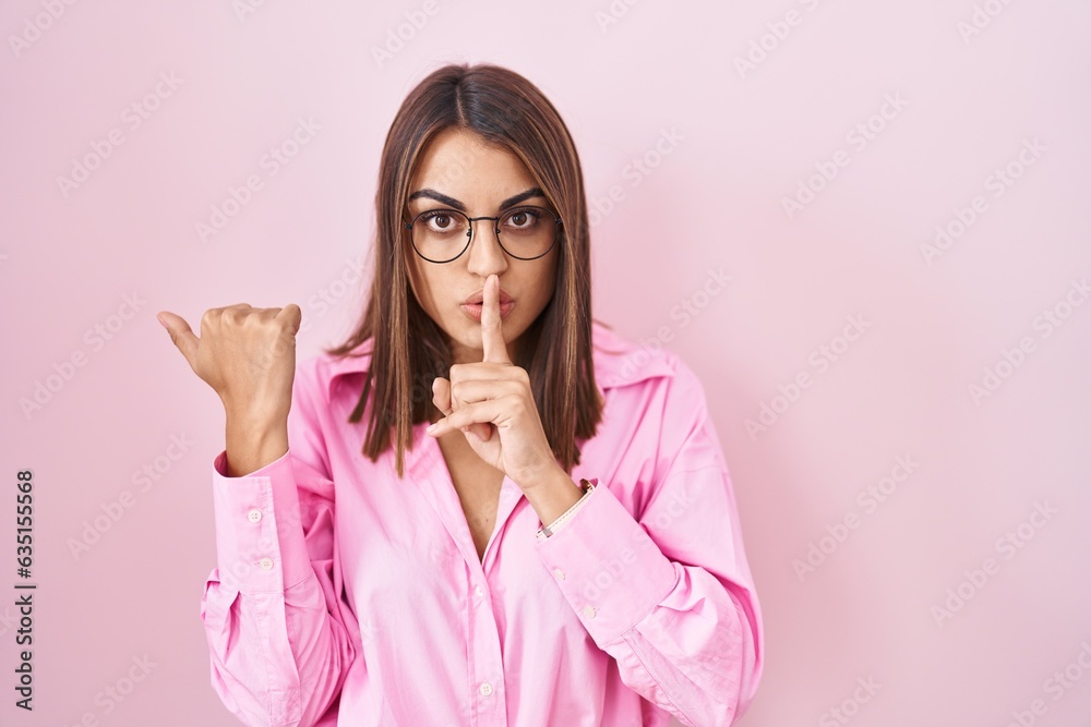 Young hispanic woman wearing glasses standing over pink background asking to be quiet with finger on lips pointing with hand to the side. silence and secret concept. Young hispanic woman wearing glasses standing over pink background asking to be quiet with finger on lips pointing with hand to the side. silence and secret concept.