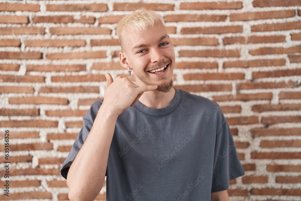 Young caucasian man standing over bricks wall smiling doing phone gesture with hand and fingers like talking on the telephone. communicating concepts. Young caucasian man standing over bricks wall smiling doing phone gesture with hand and fingers like talking on the telephone. communicating concepts.