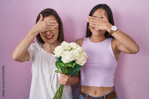 Hispanic mother and daughter holding bouquet of white flowers smiling and laughing with hand on face covering eyes for surprise. blind concept.