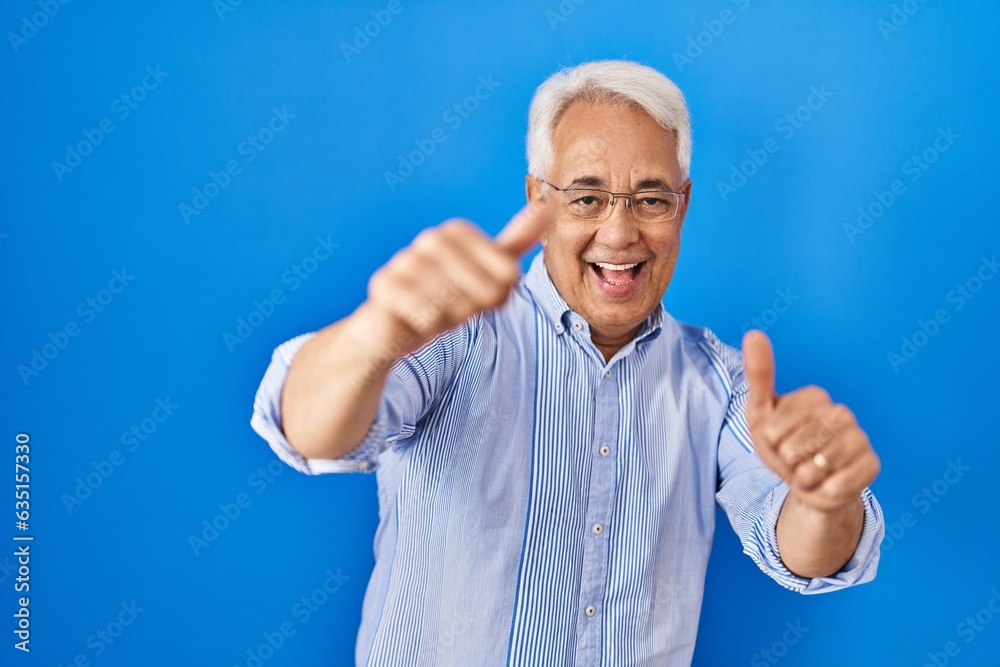 Hispanic senior man wearing glasses approving doing positive gesture with hand, thumbs up smiling and happy for success. winner gesture. Hispanic senior man wearing glasses approving doing positive gesture with hand, thumbs up smiling and happy for success. winner gesture.