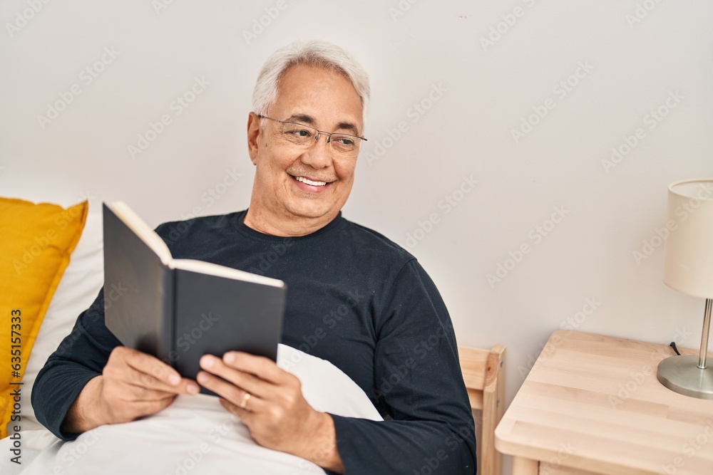Senior man reading book sitting on bed at bedroom