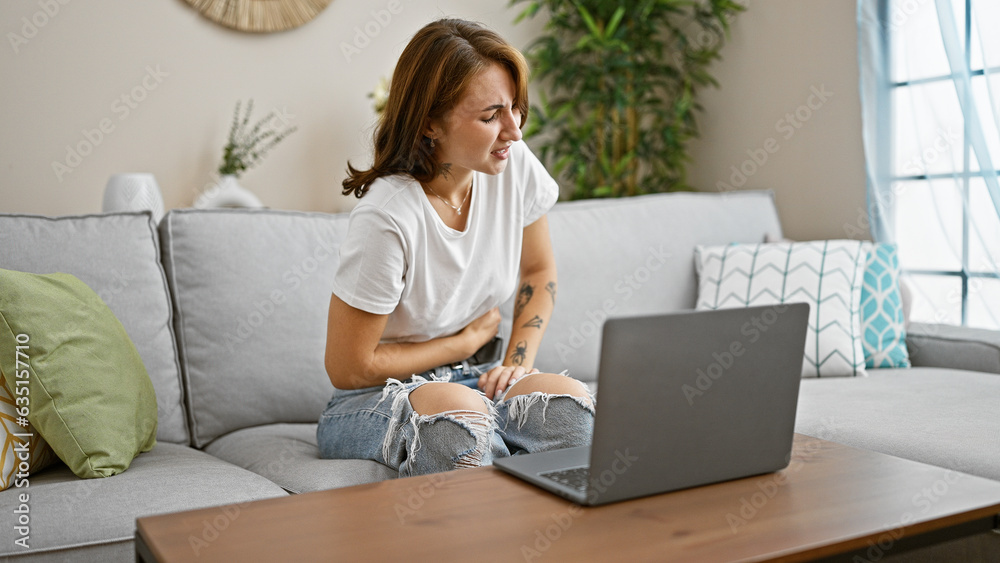 Young woman using laptop sitting on sofa suffering for stomachache at home Young woman using laptop sitting on sofa suffering for stomachache at home