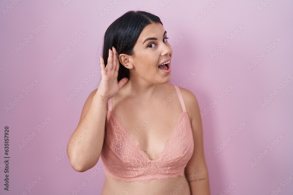 Young hispanic woman wearing pink bra smiling with hand over ear listening an hearing to rumor or gossip. deafness concept. Young hispanic woman wearing pink bra smiling with hand over ear listening an hearing to rumor or gossip. deafness concept.