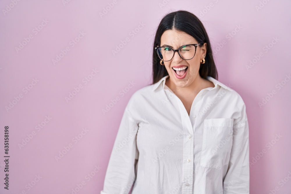 Young brunette woman standing over pink background winking looking at the camera with sexy expression, cheerful and happy face.