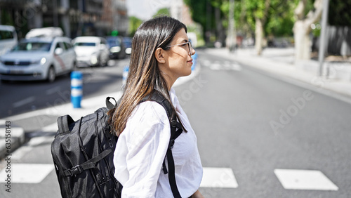 Young beautiful hispanic woman crossing crosswalk wearing a backpack in the streets of Madrid