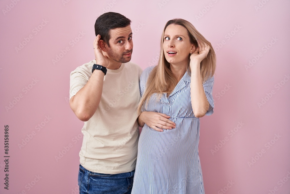 Fototapeta premium Young couple expecting a baby standing over pink background smiling with hand over ear listening an hearing to rumor or gossip. deafness concept.