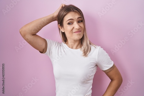 Blonde caucasian woman standing over pink background confuse and wondering about question. uncertain with doubt  thinking with hand on head. pensive concept.