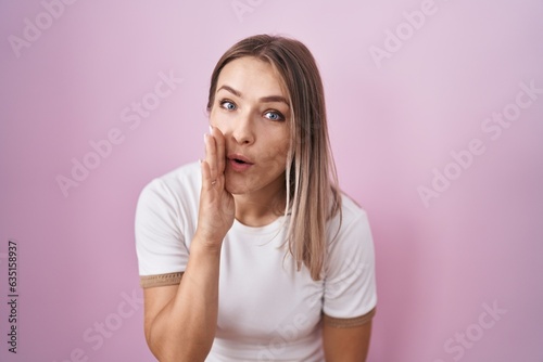 Blonde caucasian woman standing over pink background hand on mouth telling secret rumor, whispering malicious talk conversation