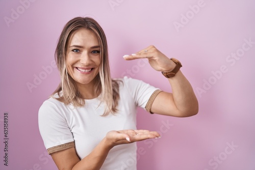 Blonde caucasian woman standing over pink background gesturing with hands showing big and large size sign, measure symbol. smiling looking at the camera. measuring concept.