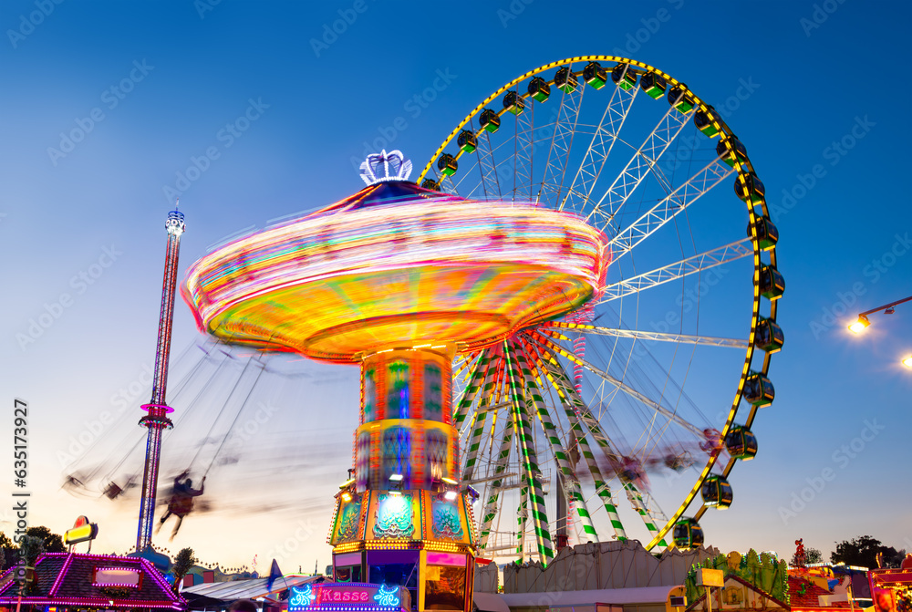 Funfair panorama wiht ferris wheel and carousels on big german funfair or Kirmes at blue hour twilight. Long time exposure with multi color light traces. - obrazy, fototapety, plakaty 