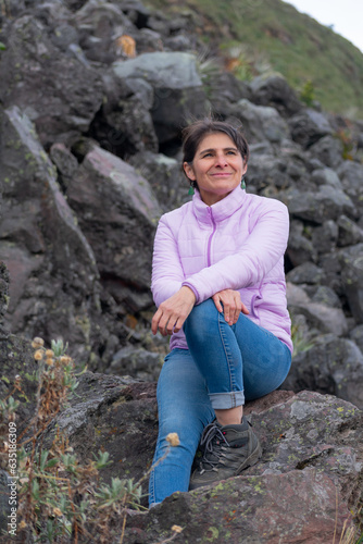 Smiling mature latin hiker woman sitting on a rock in the mountain enjoying a trekking day. Carefree climbing healthy lifestyle.