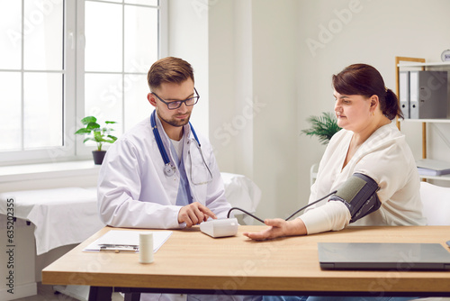 Doctor examines female patient with hypertension. Man physician in white medical coat sitting at desk with fat overweight young woman and using sphygmomanometer to measure her arterial blood pressure