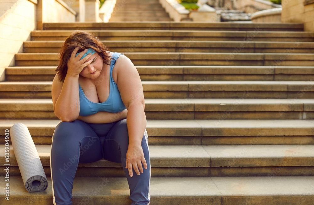 Obraz premium Tired chubby overweight woman in sportswear with fat belly and fitness mat sitting on stone stairway steps outside gym, holding hand on head, feeling stressed, sad and exhausted. Sport fatigue concept
