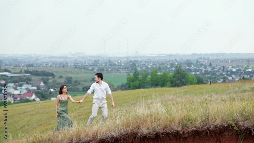 Positive man walks with woman by hand up hilly field in cloudy weather. View of big village and walk in countryside concept slow motion Positive man walks with woman by hand up hilly field in cloudy weather. View of big village and walk in countryside concept slow motion