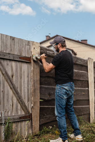A man with a powerful tool with an angle grinder cleans the wood of the fence - grinding the boards with a coarse grinder brush