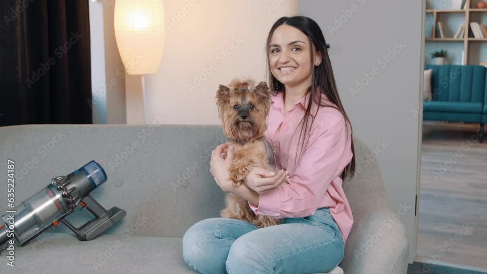 Happy young adult girl sitting on cozy sofa enjoying playing with small cute pet dog in apartment. Cordless vacuum cleaner nearby Happy young adult girl sitting on cozy sofa enjoying playing with small cute pet dog in apartment. Cordless vacuum cleaner nearby