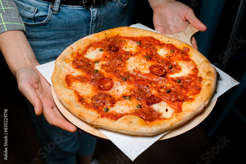 Waiter holds wooden serving board with fresh baked spicy pizza with chilli peppers, sausage, green peas and cheese