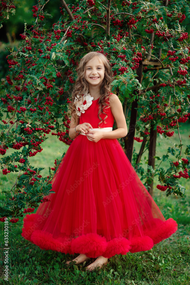 Little cheerful girl in long red dress standing near tree with red berries and laughing in green summer park, vertical outdoor colorful emotional kid's full length portrait, idea of happy childhood 