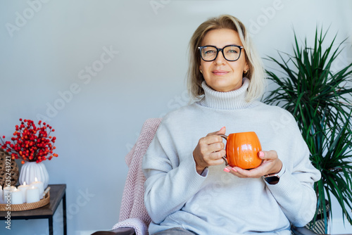 Middle aged woman relaxing with pumpkin shaped cup of hot drink in scandy style hygge interior home with fall mood decor. Lady dreaming, enjoy calm mood without stress, well being alone. Cozy autumn. photo