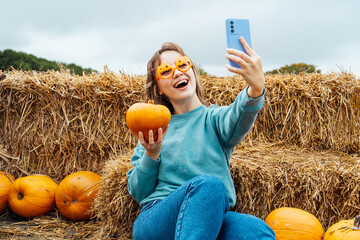 Smiling woman in fun glasses sitting on straw bales and holding pumpkin and making selfie on phone. Selecting Thanksgiving and Halloween holidays decor on agriculture farm. Autumn fall festive mood.